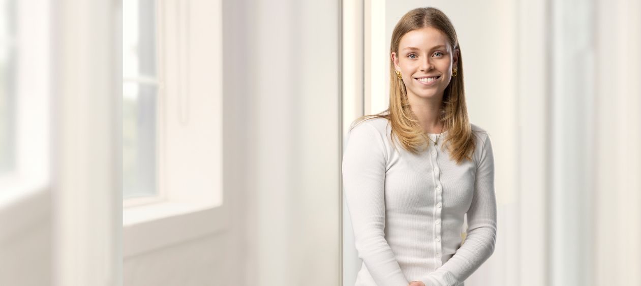 Smiling woman in a white long-sleeve top standing in a bright, sunlit hallway by a window.